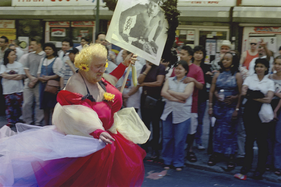 thumbnail of Photo of an elderly man in a red dress holding up a sign of a man, running through the streets