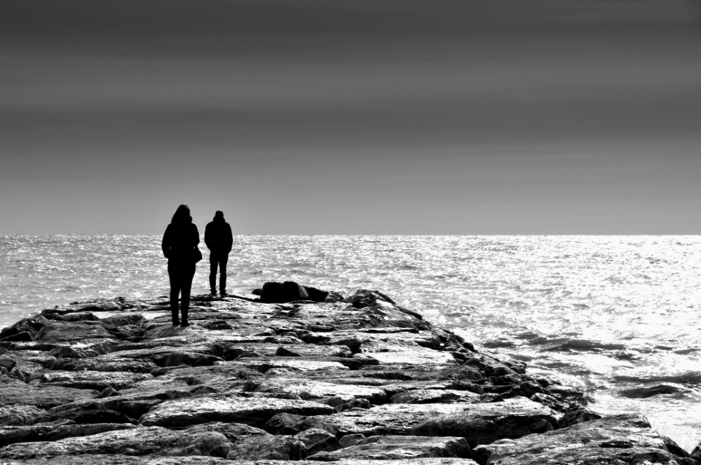 thumbnail of Photograph of two silhouettes standing on rocks in front of the water